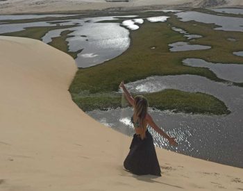 Mulher abrindo os braços com a vista das dunas e lagoa ao fundo